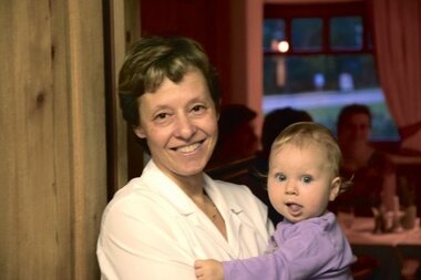 Eine Frau lächelt neben einer Wand, während sie ein kleines Kind im Arm hält.