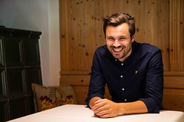 Ein Mann mit kurzen Haaren, der in einem Holzraum an einem Tisch sitzt und lächelt.
