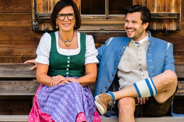 Ein Paar in traditioneller bayerischer Tracht sitzt lächelnd auf einer Holzbank vor einer Holzwand.