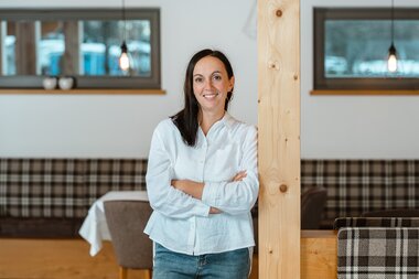 Eine lächelnde Frau in einem weißen Hemd steht mit verschränkten Armen neben einem Holzpfeiler in einem modernen Café-Inneren.
