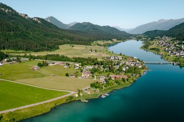 Aerial view of a tranquil lake surrounded by green fields and mountains, with small houses along the shore.
