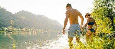 A couple in swimwear walks hand-in-hand along the shore of a lake.