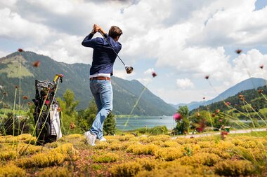 A golfer swings a club on a grassy area, with mountains and a lake in the background under a cloudy sky.