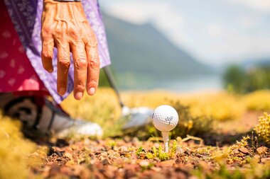 Eine Person in traditioneller Kleidung bereitet sich darauf vor, einen Golfball auf einem Tee zu schlagen, umgeben von Natur.