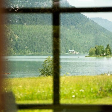 Blick auf einen ruhigen See, umrahmt von Fensterrahmen, umgeben von üppigen grünen Bergen und Bäumen.