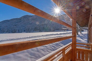 View from a balcony overlooking a snowy landscape with mountains in the background and the sun shining through trees.
