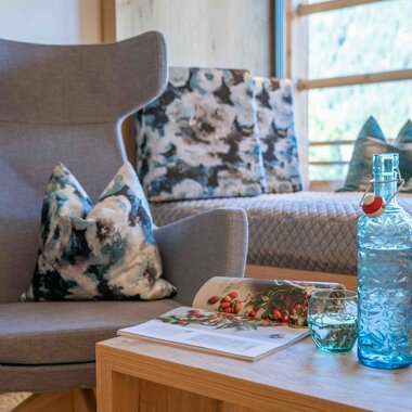 A modern chair with cushions, a wooden table with a magazine and glasses, and a bed with colorful pillows in the background.