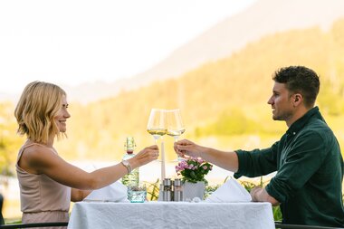 A couple toasting with wine glasses at an outdoor dinner setting, with scenic mountains in the background.