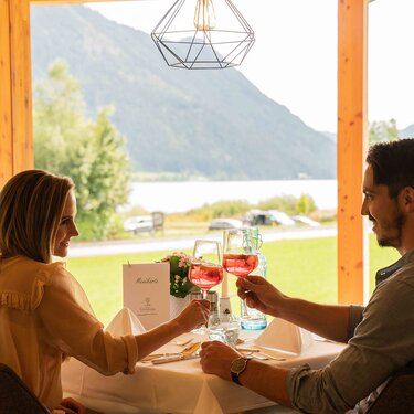 Ein Paar sitzt an einem Tisch mit Blick auf den See und prostet mit Getränken.
