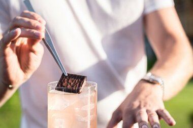 Ein Barkeeper fügt mit einer Pinzette ein Schokoladenstückchen auf einen Cocktail in einem Glas mit Eis hinzu.