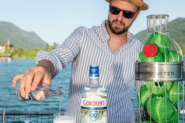 Mann mit Sonnenbrille, der ein Getränk mit Gordon's Gin an einem Steg am Wasser zubereitet.