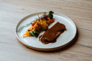 Ein kunstvoll angerichtetes Gericht mit einem Stück gegrilltem Fleisch in reicher Sauce, begleitet von buntem sautiertem Gemüse auf einem runden weißen Teller.