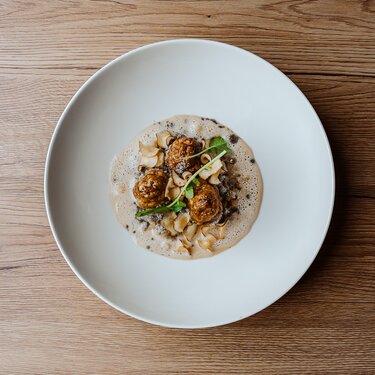 A beautifully plated dish on a white plate featuring three round, fried dumplings, surrounded by a creamy sauce and garnished with fine herbs.