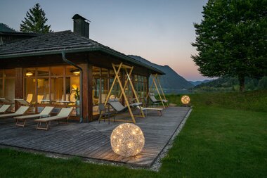 Modern wooden deck with seating and decorative lights, surrounded by mountains and grass.