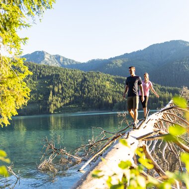A man and woman walking along a fallen tree by a serene lake surrounded by mountains and greenery.