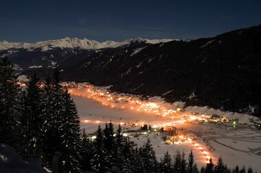 Night view of a snow-covered valley with illuminated houses and mountains in the background.