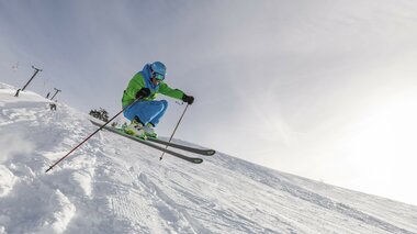 Ein Skifahrer in blau-grüner Kleidung springt über schneebedecktes Gelände vor einem hellen Himmel.