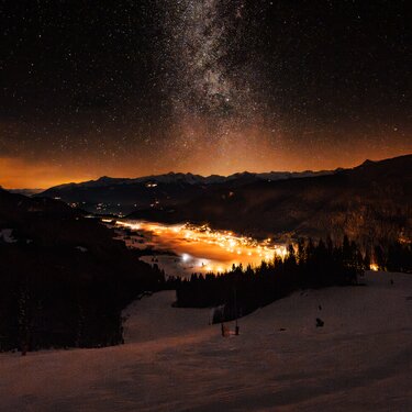 A breathtaking night sky filled with stars over a snow-covered valley with soft glowing lights from a nearby town.