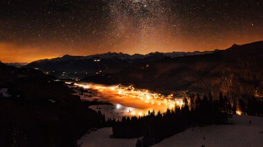 Ein atemberaubender Nachthimmel voller Sterne über einem schneebedeckten Tal mit sanften Lichtern einer nahegelegenen Stadt.