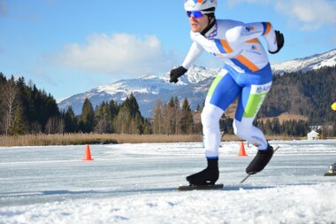 A speed skater in a blue and white suit races on an outdoor ice rink surrounded by mountains.