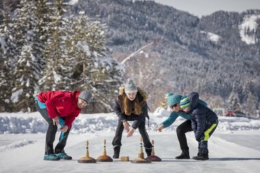 Three people playing curling on an outdoor ice rink surrounded by snow-covered trees and mountains.