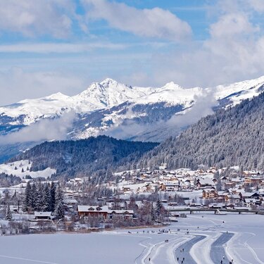 A winter landscape featuring snow-covered mountains and a village under a blue sky with fluffy clouds.