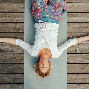Eine Frau, die auf einer Yogamatte liegt, mit ausgestreckten Armen, in einem weißen Oberteil und buntem Rock, umgeben von Holzboden.