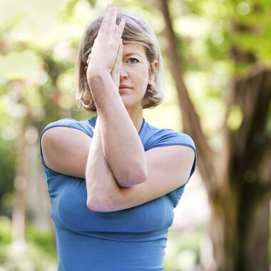A woman in a blue shirt demonstrates a stretch, with one arm crossed over her face in a green park.