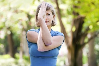 A woman in a blue shirt demonstrates a stretch, with one arm crossed over her face in a green park.
