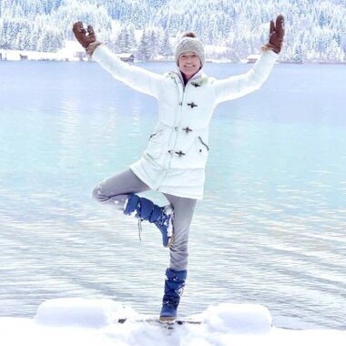 A person standing on one leg by a snowy lake in winter, wearing a white coat and a knitted hat.