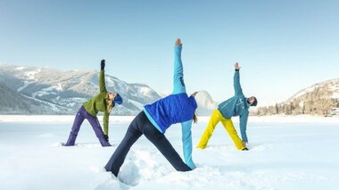 Drei Personen machen im Schnee eine Dehnübung vor einer Berglandschaft.