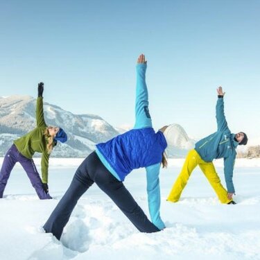 Drei Personen in Winterkleidung machen Yoga-Übungen im Schnee mit Bergen im Hintergrund.